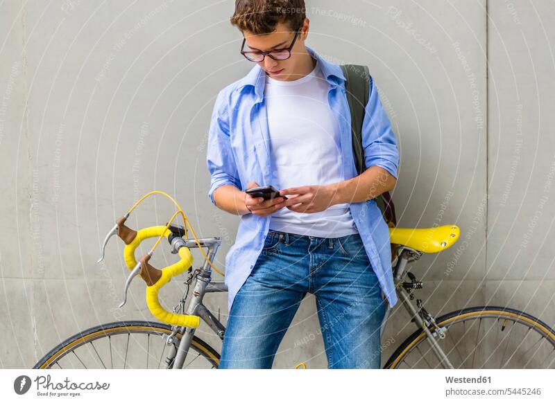 Young man with racing cycle looking at cell phone in front of concrete wall men males Adults grown-ups grownups adult people persons human being humans