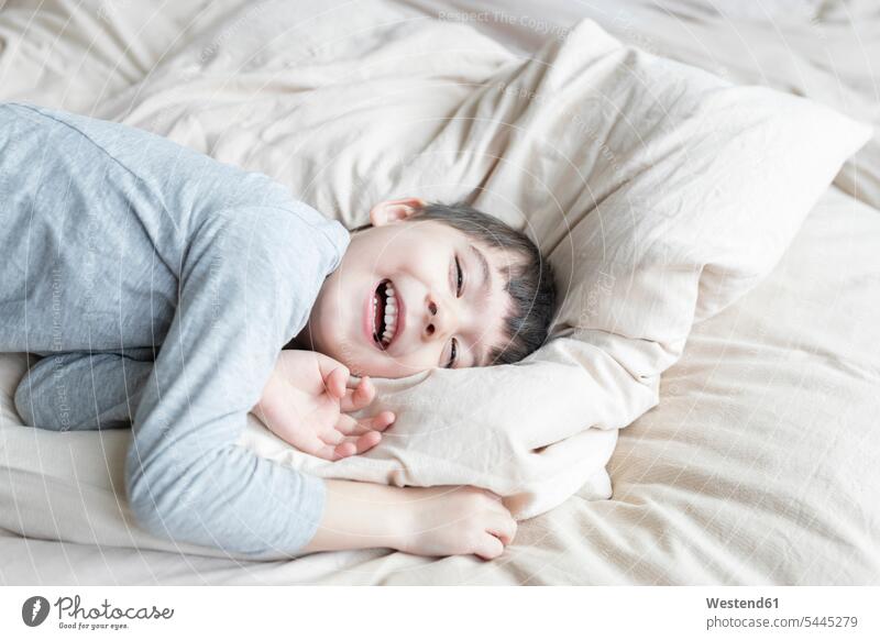 Happy boy lying in bed playing boys males laughing Laughter cushion cushions beds laying down lie lying down child children kid kids people persons human being