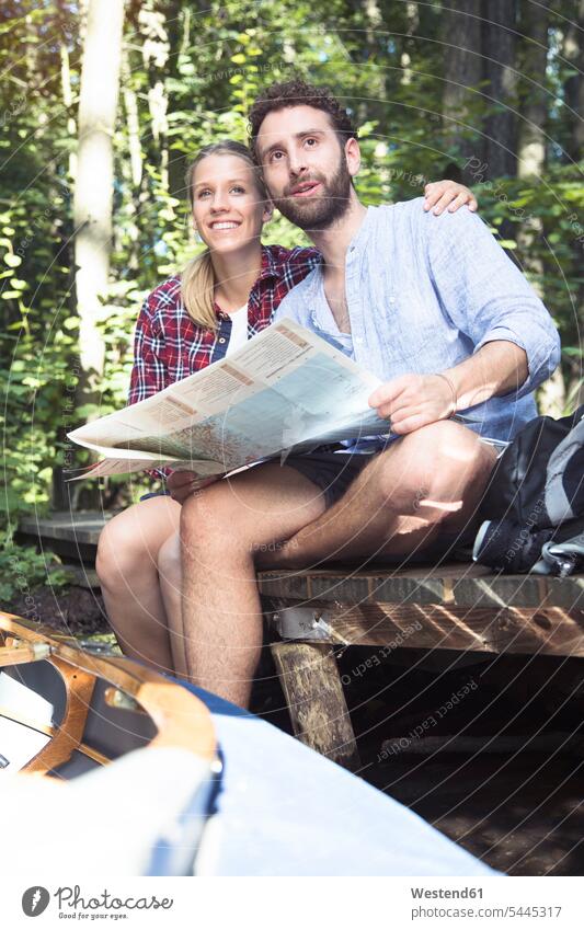 Young couple with map and canoe sitting on a jetty at a forest brook woods forests twosomes partnership couples brooks rivulet Seated jetties maps canoes boat