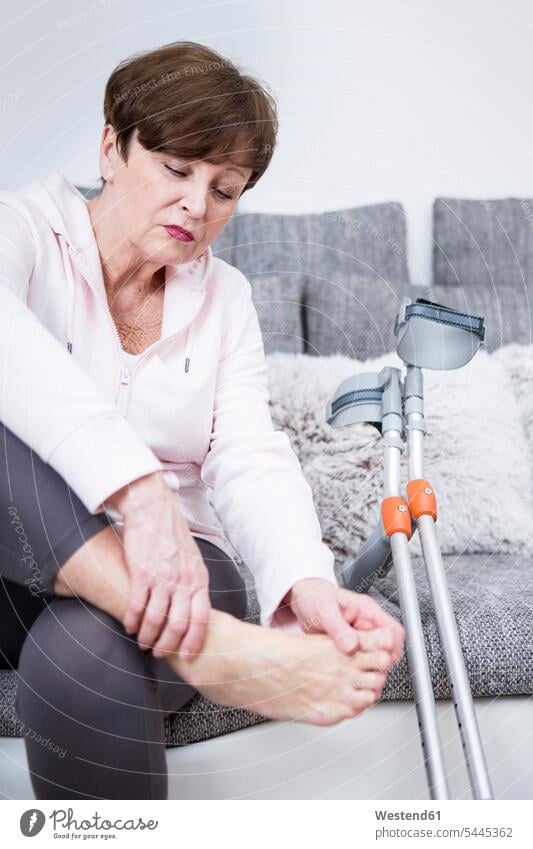 Senior woman with crutches sitting on couch, checking her ankle watching looking looking at foot human foot human feet pain females women senior women