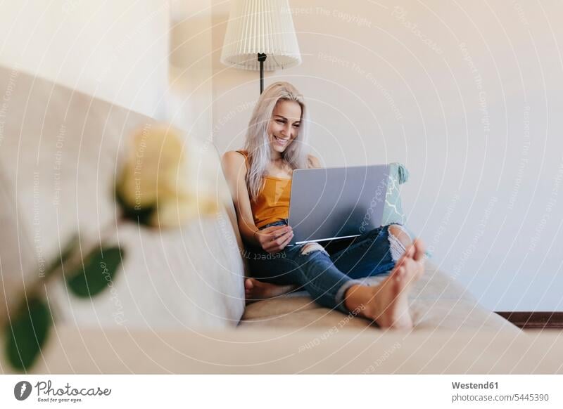 Smiling young woman using laptop on couch at home smiling smile settee sofa sofas couches settees Laptop Computers laptops notebook females women computer