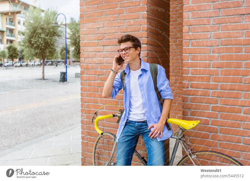 Portrait of smiling young man with racing cycle on the phone standing in front of brick wall men males call telephoning On The Telephone calling Adults