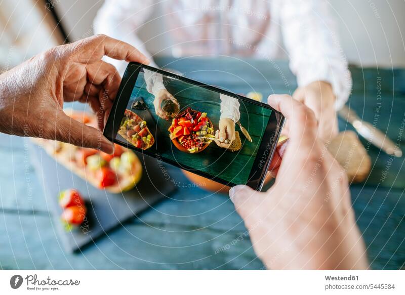 Close-up of man's hands taking a picture with mobile phone eating salad of tomato, pomegranate, papaya and olives, with papaya with fruits on the side and with glass of wine