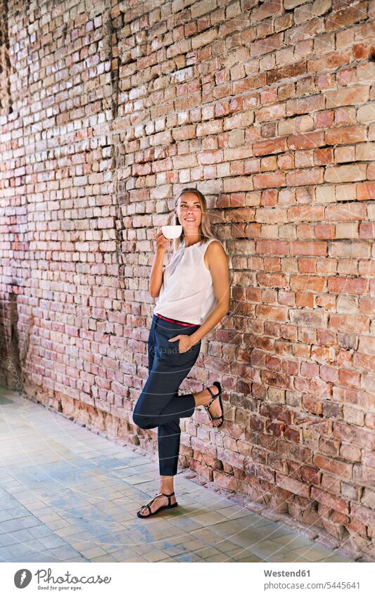 Businesswoman drinking cup of coffee at brick wall in office Coffee females women businesswoman businesswomen business woman business women smiling smile Drink