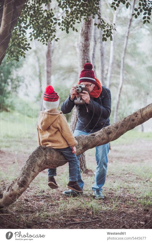 Father taking a picture of his son sitting on tree trunk in forest photographing boy boys males father pa fathers daddy dads papa sons manchild manchildren kid