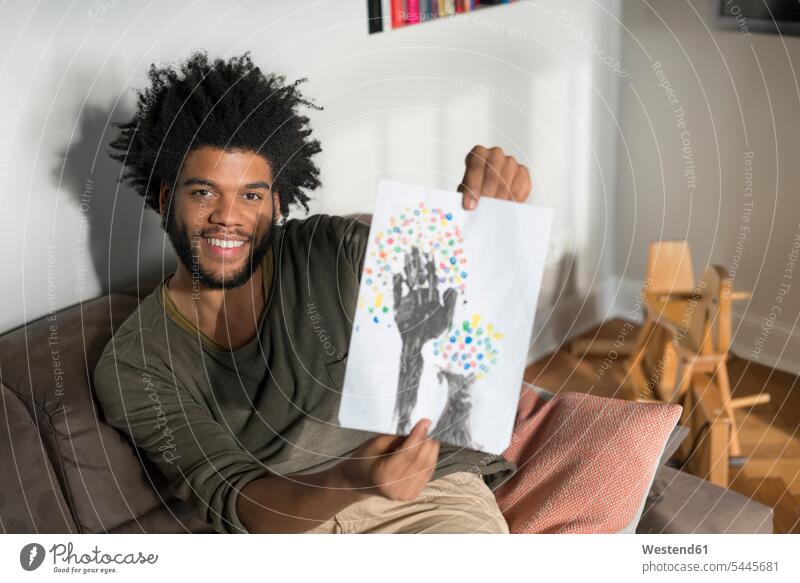 Man sitting on couch in living room showing children's drawing smiling smile man men males Seated child's drawing Child's Drawings Adults grown-ups grownups