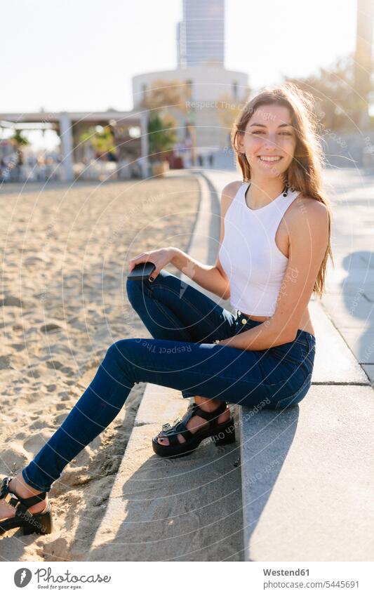 Spain, Barcelona, portrait of smiling young woman sitting on beach promenade at sunset sea front boardwalk portraits females women promenades Adults grown-ups