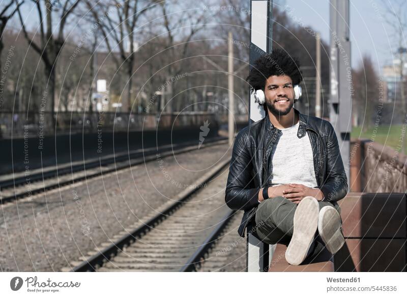 Smiling man sitting on wall listening to music next to train rails men males smiling smile Seated headphones headset Adults grown-ups grownups adult people