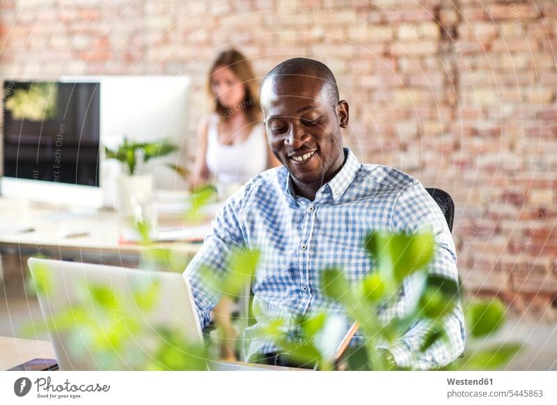 Smiling businessman using laptop at desk in office with colleague in background desks Laptop Computers laptops notebook use offices office room office rooms