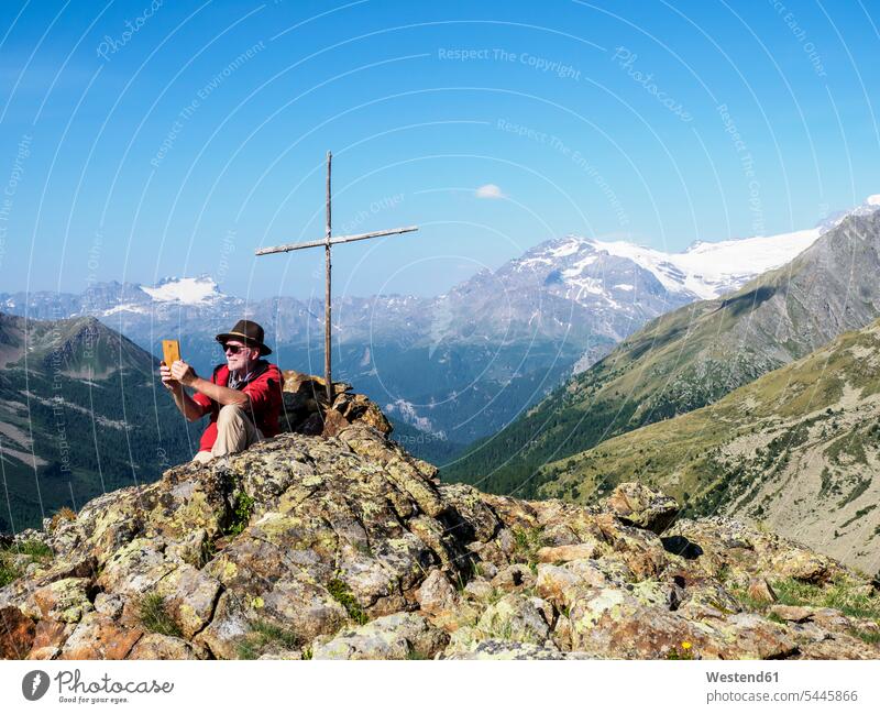 Italy Lombardy, Passo di Val Viola, Man taking pictures with his smartphone, sitting at summit cross summit crosses Seated mountain range mountains