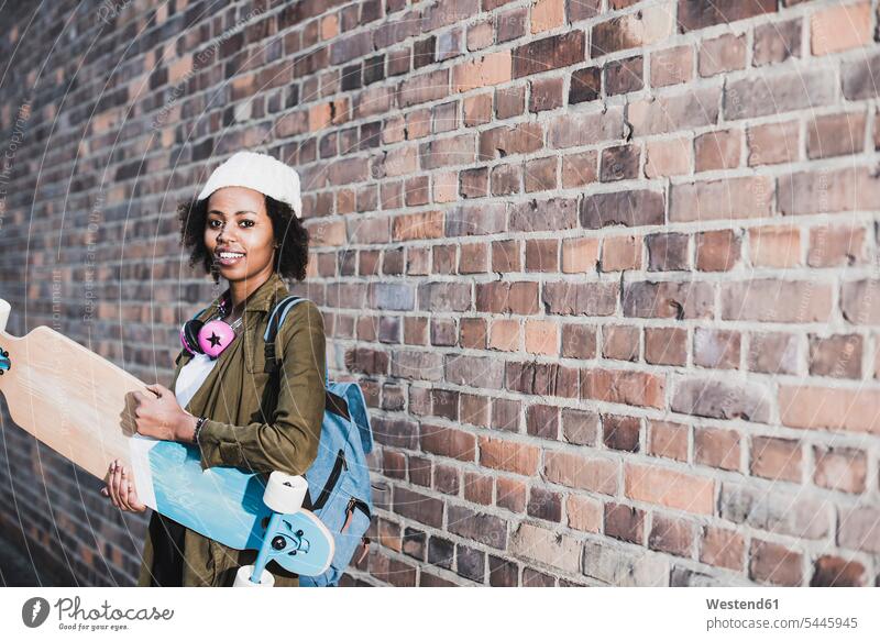 Portrait of smiling young woman with headphones, skateboard and backpack in front of brick wall Skate Board skateboards portrait portraits females women Adults