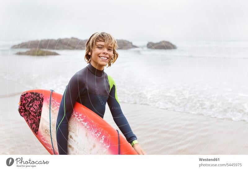 Spain, Aviles, portrait of smiling young surfer carrying surfboard on the beach surfers surfboards beaches sea ocean smile Teenager Teens teenagers surfing