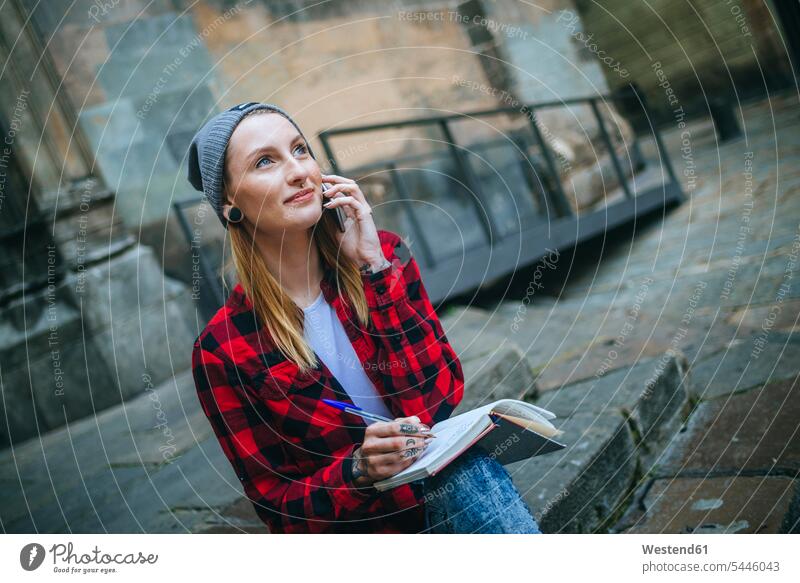 Spain, Barcelona, portrait of smiling young woman on the phone sitting on stairs with notebook females women notebooks call telephoning On The Telephone calling