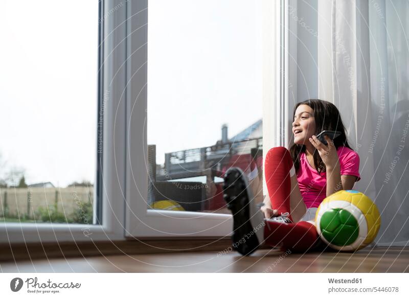 Girl in soccer outfit sitting on floor in living room speaking to someone on her smartphone soccer ball soccer balls footballs girl females girls mobile phone