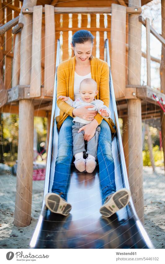 Happy baby girl sitting with her mother on shute on playground infants nurselings babies slide Chute playground slide slides Chutes Seated play yard play ground