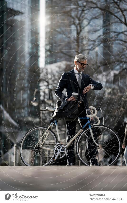Grey-haired businessman with bicycle and coffee to go in the city looking at smartwatch Businessman Business man Businessmen Business men town cities towns