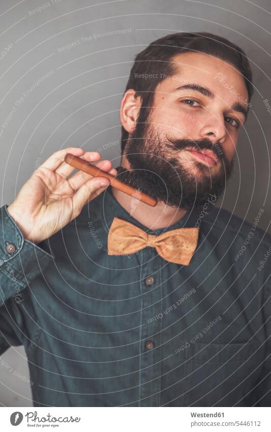 Man combing his beard with a wooden brush, wearing denim shirt and cork bow tie portrait portraits man men males people persons human being humans human beings