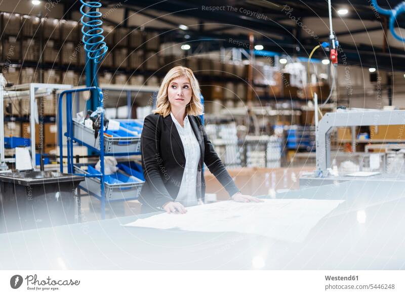 Blond businesswoman standing on shop floor, looking at plans manager female managers production hall eyeing businesswomen business woman business women blond