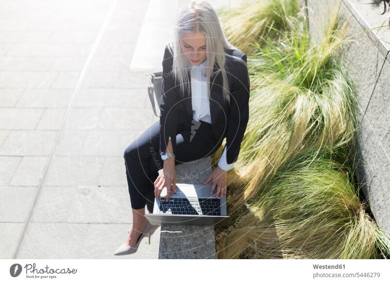 Young businesswoman sitting on a wall in the city using laptop businesswomen business woman business women females Laptop Computers laptops notebook Seated