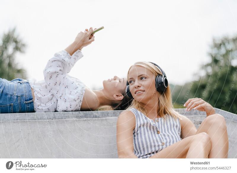 Two young women with cell phone and headphones in a skatepark Skateboard Park skate park female friends headset mobile phone mobiles mobile phones Cellphone
