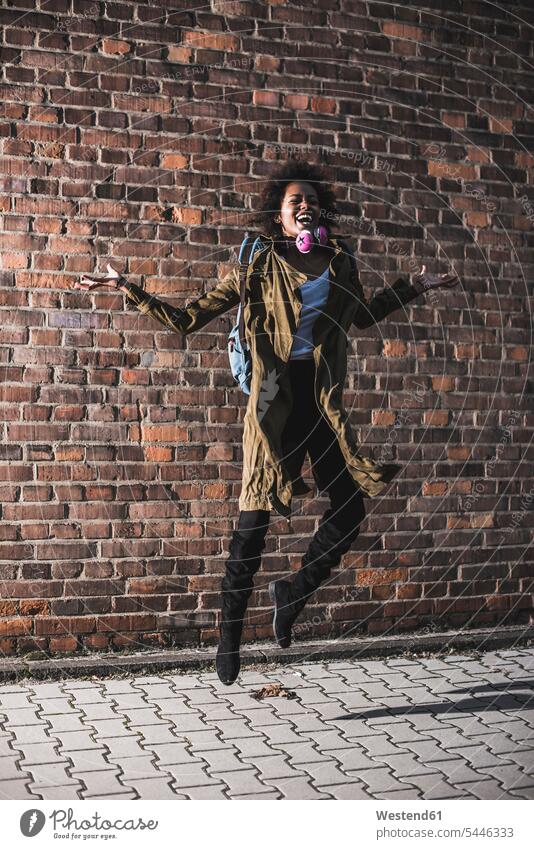 Happy young woman jumping in the air in front of brick wall females women Leaping jump in the air Adults grown-ups grownups adult people persons human being