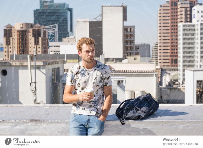 Young man standing on rooftop terrace drinking coffee men males enjoying indulgence enjoyment savoring indulging blond blond hair blonde hair roof terrace deck