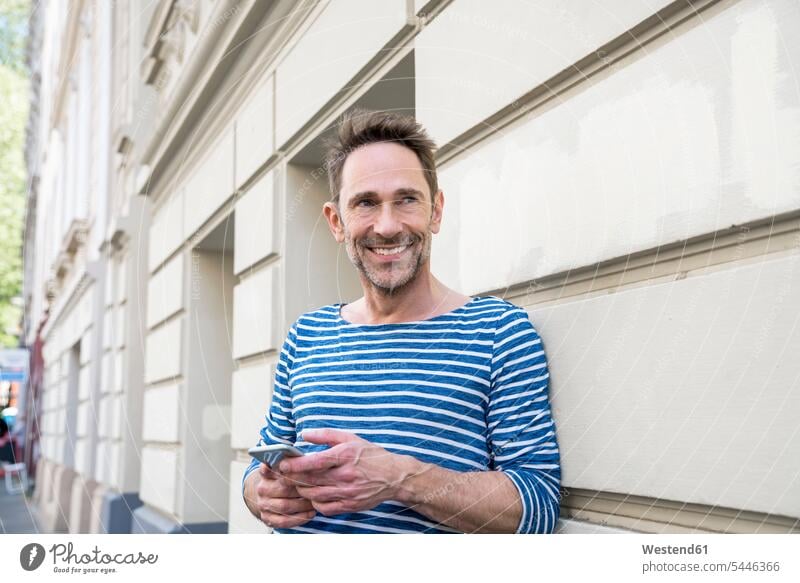 Portrait of smiling with smartphone leaning against facade portrait portraits man men males Adults grown-ups grownups adult people persons human being humans