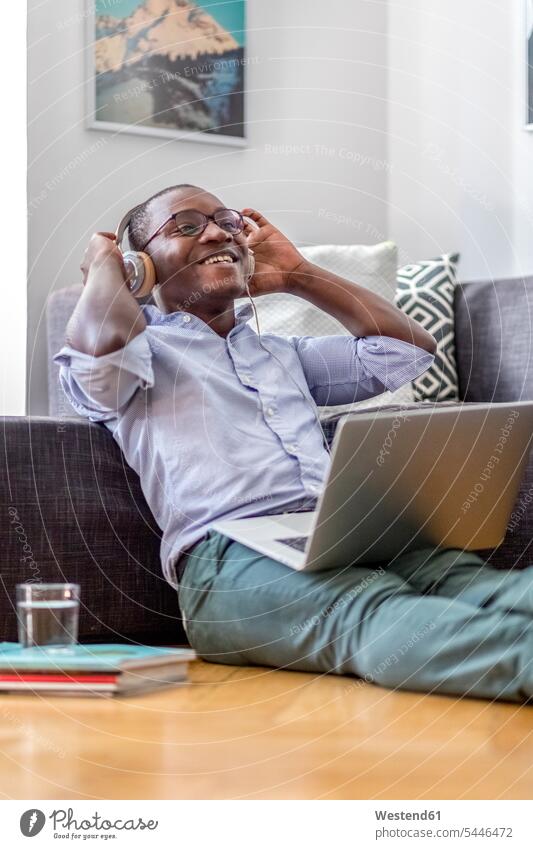Happy young man sitting on the floor in the living room using laptop and headphones men males portrait portraits headset Adults grown-ups grownups adult people