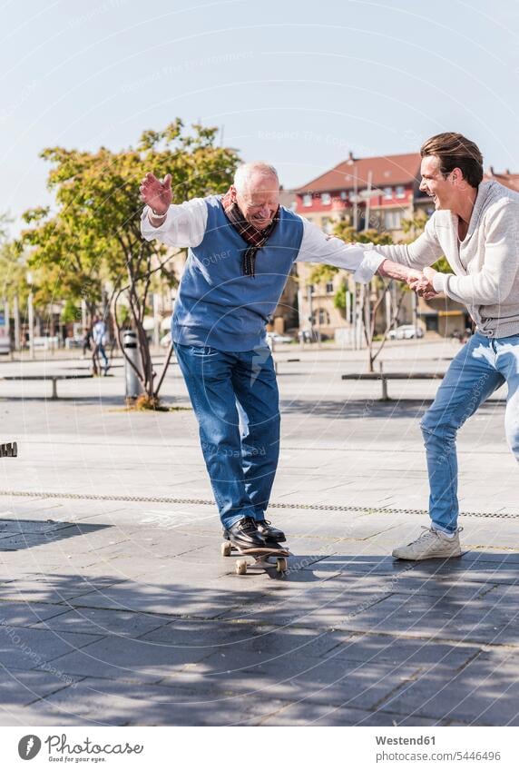 Adult grandson assisting senior man on skateboard grandfather grandpas granddads grandfathers Skate Board skateboards happiness happy grandsons grandparents