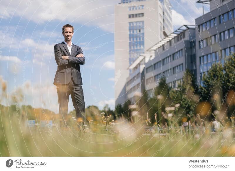 Businessman standing on field in front of office buildings Business man Businessmen Business men meadow meadows built structure built structures business people