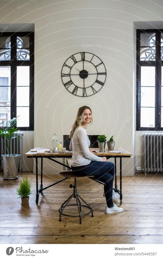 Portrait of smiling woman sitting at desk at home under large wall clock desks wall clocks portrait portraits females women Seated smile Table Tables Clock