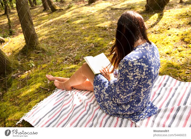 Young woman sitting on blanket in forest reading a book Seated females women woods forests books relaxed relaxation Adults grown-ups grownups adult people