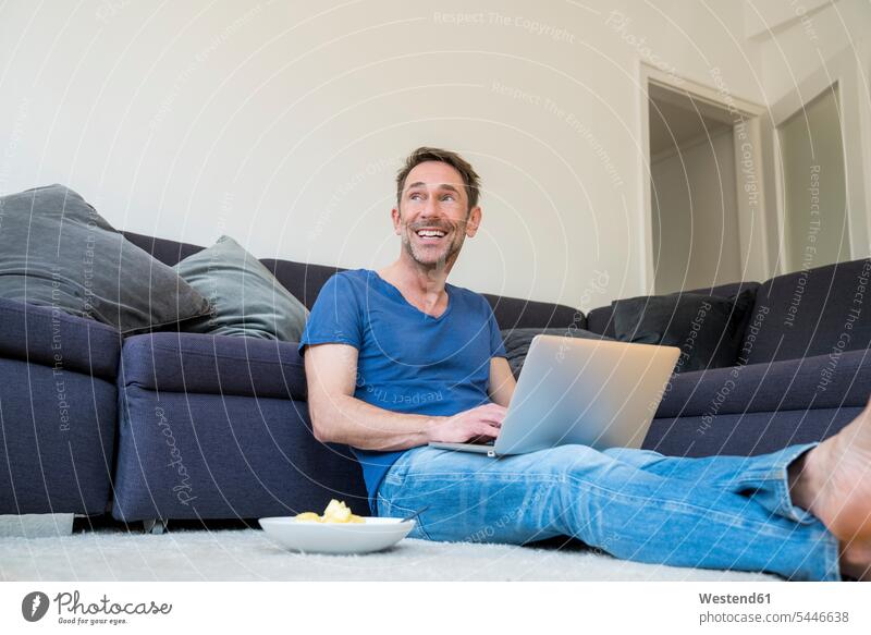 Portrait of laughing man with laptop sitting on the floor in the living room men males Laptop Computers laptops notebook Adults grown-ups grownups adult people