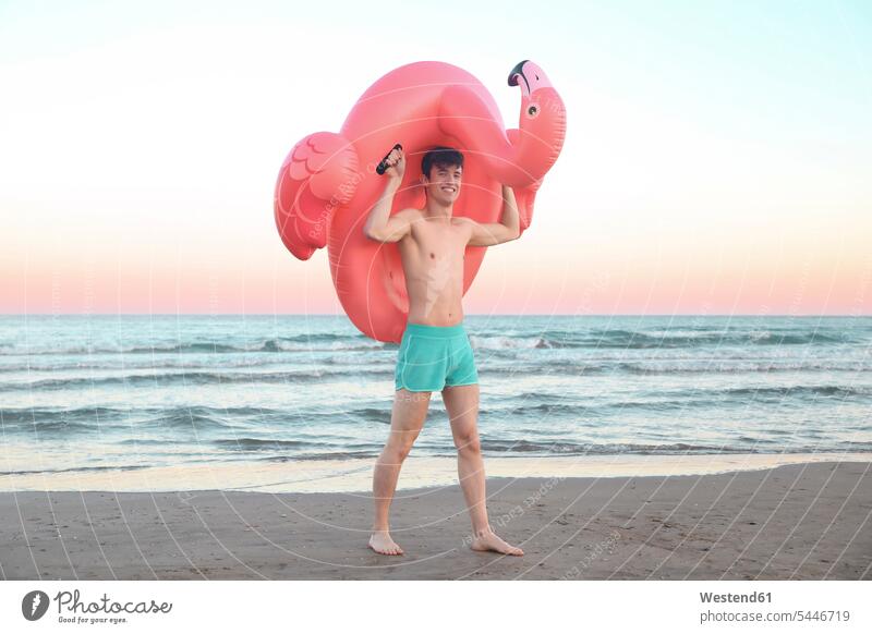 Smiling young man with inflatable pink flamingo on the beach at sunset beaches men males Adults grown-ups grownups adult people persons human being humans