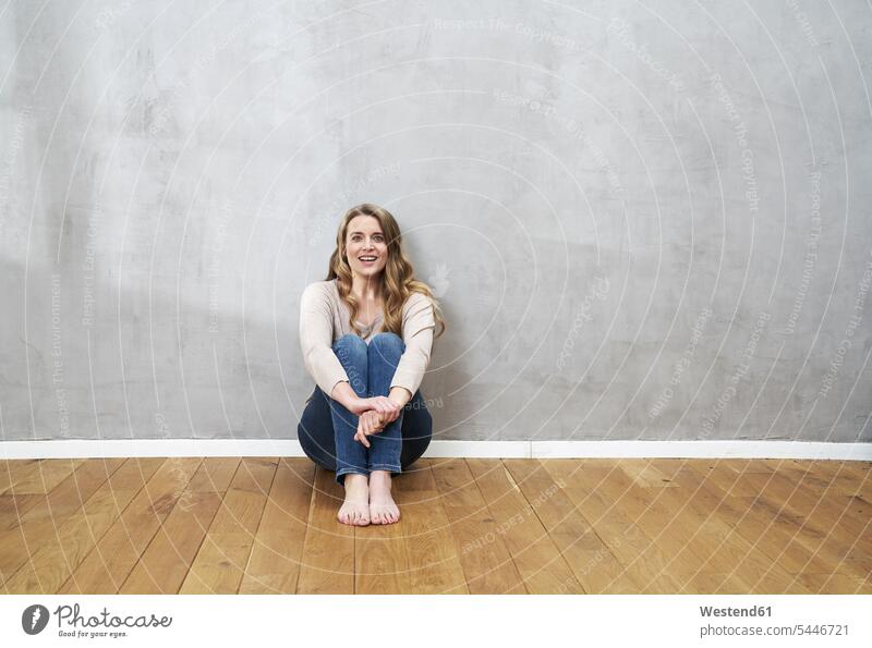 Smiling blond woman sitting on the floor in front of grey wall portrait portraits females women Adults grown-ups grownups adult people persons human being