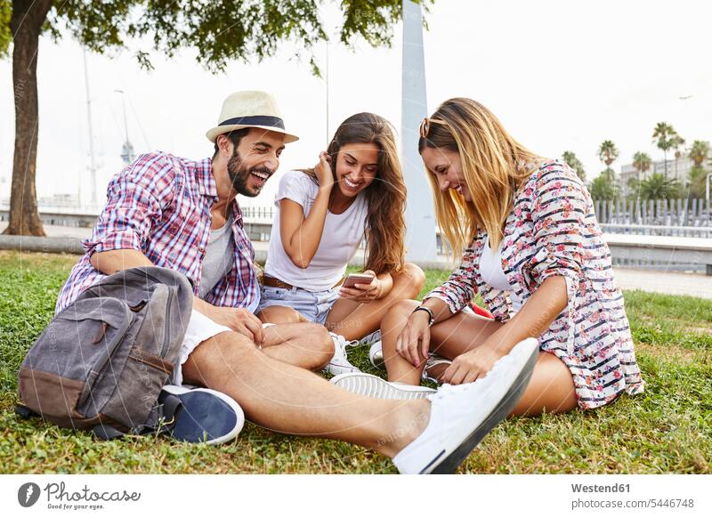 Three happy friends sitting in park sharing smartphone mate laughing Laughter mobile phone mobiles mobile phones Cellphone cell phone cell phones friendship