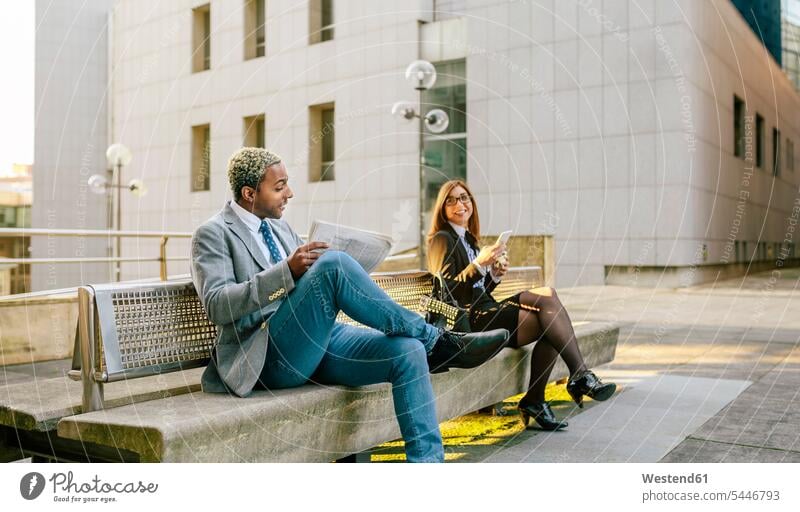 Young businessman and woman sitting on bench, talking business people businesspeople on the move on the way on the go on the road colleagues speaking newspaper