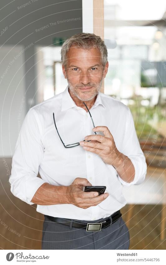 Confident businessman with cell phone in office mobile phone mobiles mobile phones Cellphone cell phones offices office room office rooms Businessman