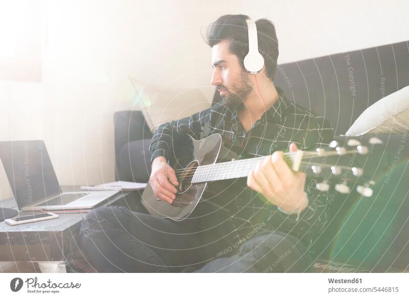 Young man at home playing guitar and wearing headphones connected to laptop men males guitars Laptop Computers laptops notebook Adults grown-ups grownups adult