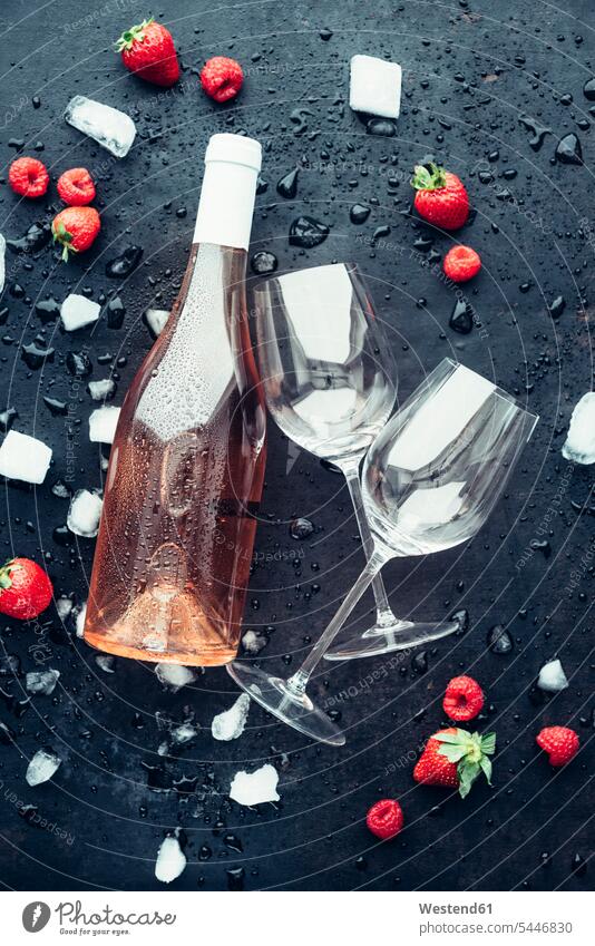 Bottle of rose wine, two empty wine glasses, icecubes, strawberries and raspberries on dark ground ice-cooled iced black background black backgrounds wet