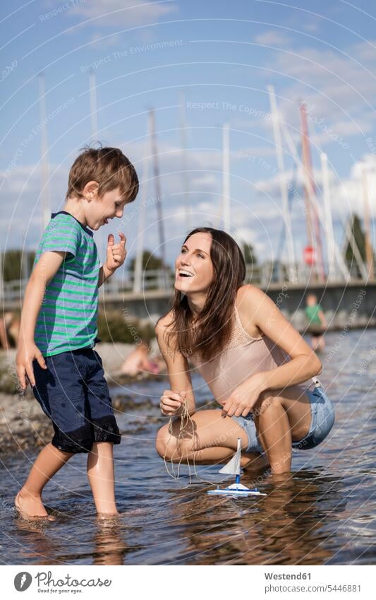 Germany, Friedrichshafen, Lake Constance, happy mother and son with toy boat at lakeshore sons manchild manchildren playing happiness looking eyeing mommy