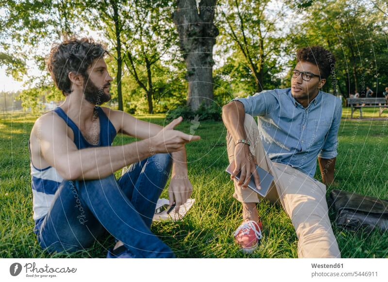 Two friends sitting in a park with mobile device and papers discussing man men males Seated parks discussion documents friendship Adults grown-ups grownups