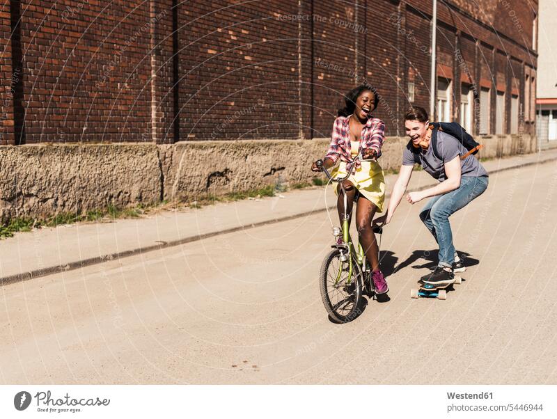 Young woman on bicycle pulling young man, standing on skateboard multicultural skateboarding Boarding Skate Board skateboards couple twosomes partnership