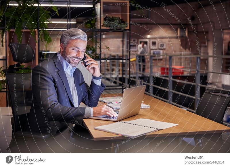 Mature businessman working in modern office, using laptop while talking on the phone sitting Seated desk desks At Work Smartphone iPhone Smartphones
