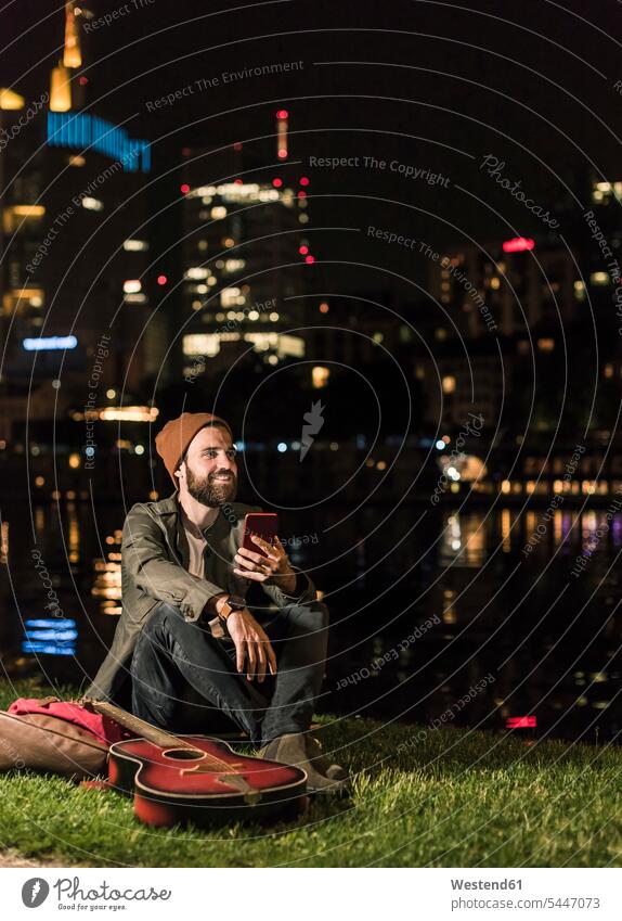 Smiling young man with guitar and cell phone sitting at urban riverside at night mobile phone mobiles mobile phones Cellphone cell phones men males smiling