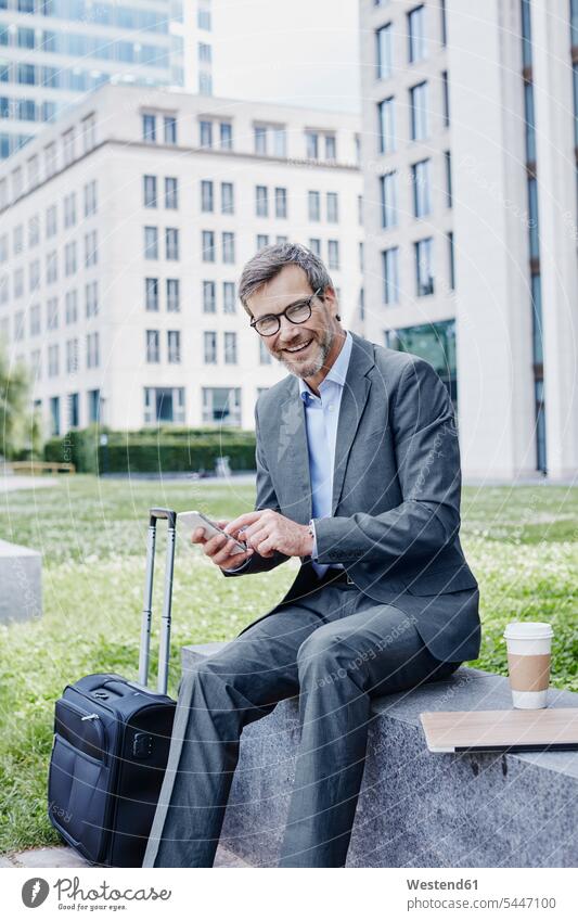 Smiling mature businessman outdoors with laptop, cell phone, takeaway coffee and rolling suitcase mobile phone mobiles mobile phones Cellphone cell phones