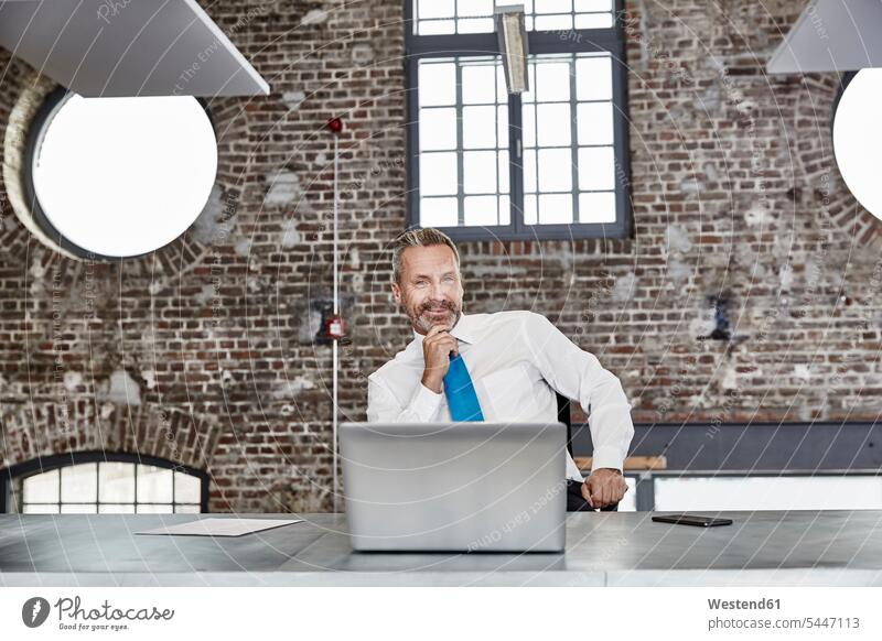 Portrait of confident businessman with laptop sitting at table in a loft Seated Laptop Computers laptops notebook smiling smile Businessman Business man