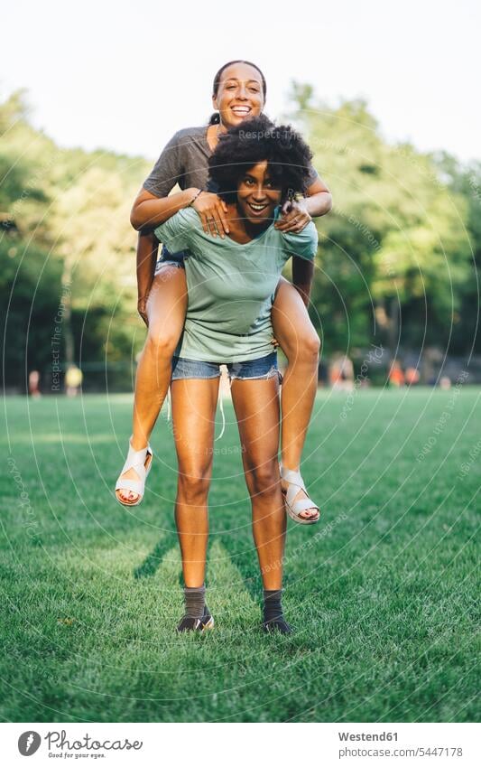 Young woman giving her friend a piggyback ride in a park piggy-back pickaback Piggybacking Piggy Back parks female friends mate friendship females women meadow