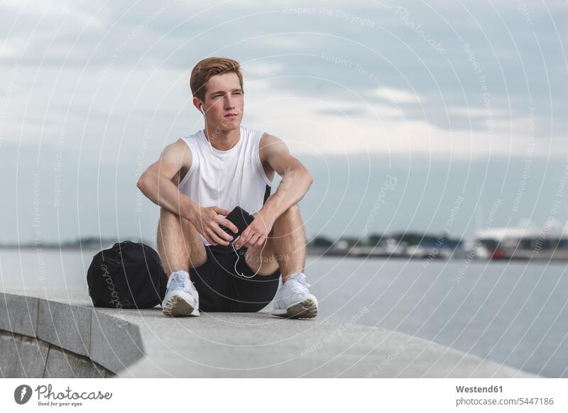 Young athlete sitting on a wall at riverbank with cell phone and earphones exercising exercise training practising Seated ear phone ear phones bag bags man men