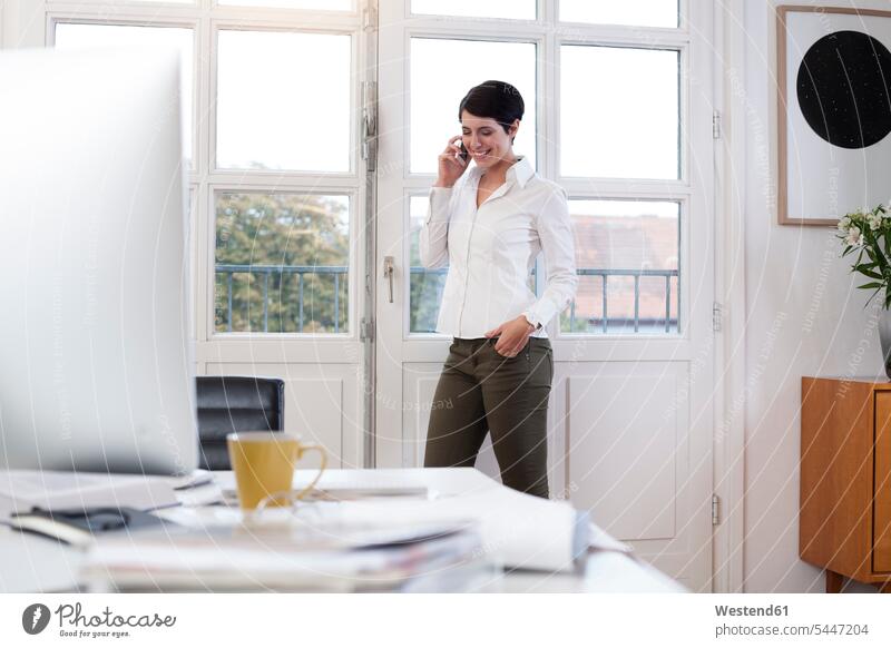 Woman standing at the window in office talking on phone smiling smile businesswoman businesswomen business woman business women on the phone call telephoning